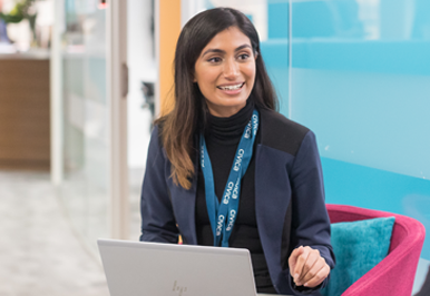 Happy cheerful millennial indian business woman sitting at desk in front of laptop laughing talking to someone in office