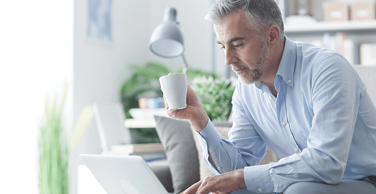 Businessman relaxing at home in the living room, he is sitting on the sofa, having a coffee and working with a laptop